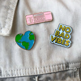 NO BAD VIBES Tekst Emaille Pin samen met een vintage roze radio pin en een hartje met planeet aarde pin op een spijkerjasje
