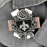 Broche patch van een wit en beige gekruist lint met daarop een zwart embleem met zilverkleurige sierknoop en franse lelie op een grijze jas
