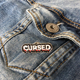 Cursed Tekst Emaille Pin op een borstzakje van een blauwe spijkerjas