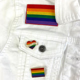Rainbow Regenboog Hartje Love Is Love Tekst Gay Pride Emaille Pin samen met een andere regenboog pin en regenboog vlag strijk patch op een wit spijkerjasje