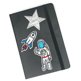Astronaut Met Thumbs Up Strijk Patch samen met een glitter ster en wit blauwe raket strijk patch op de voorkant van een zwart leren agenda
