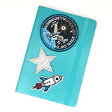 Zilver Kleurige Glitter Ster Strijk Patch samen met een blauw witte raket en een ronde space shuttle strijk patch op de voorkant van een blauwe agenda